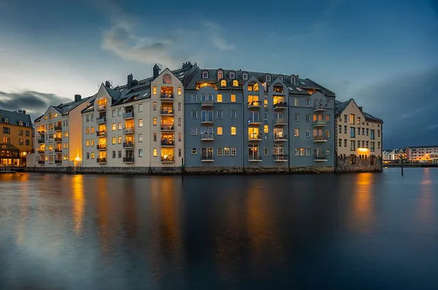 Leilighetsbygg ved vannkanten med opplyste vinduer og speilinger i vannet ved skumring.